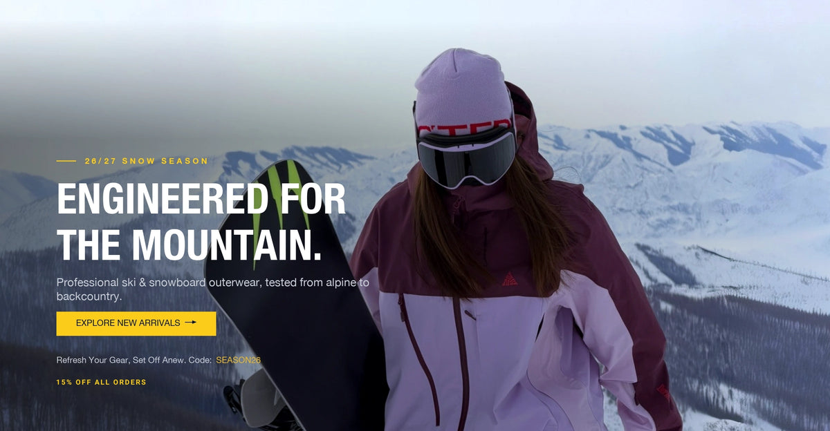 Snowboarder in a purple ski jacket and goggles holding a snowboard on a snowy mountain ridge